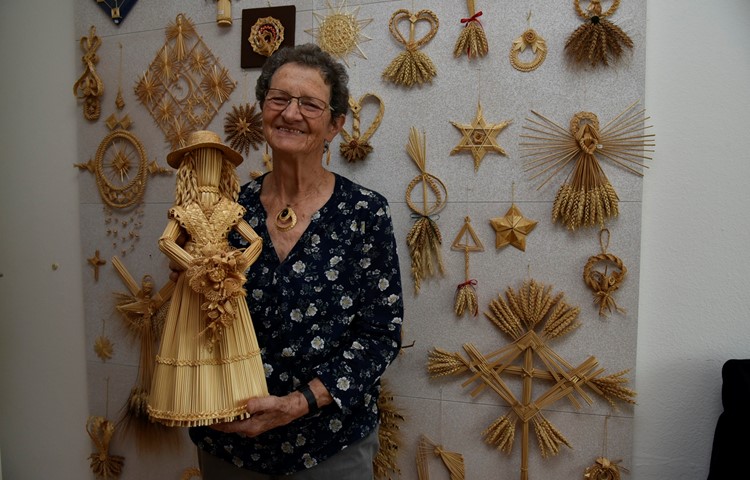 Vreni Hug vor einer Wand mit einer Auswahl ihrer Stroharbeiten. In der Hand eine Figur, deren filigranes Bouquet so viele Arbeitsstunden brauchte, wie sie für die ganze Figur benötigte.